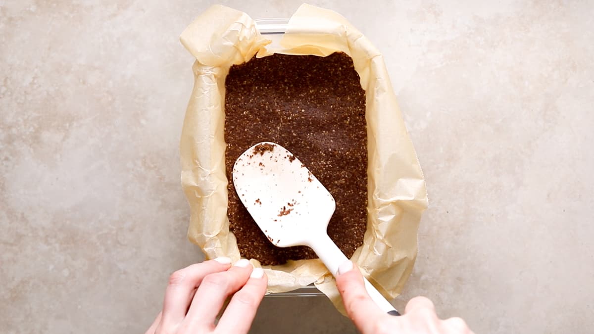 Chocolate biscuit mixture being pressed evenly into a lined container with a spatula