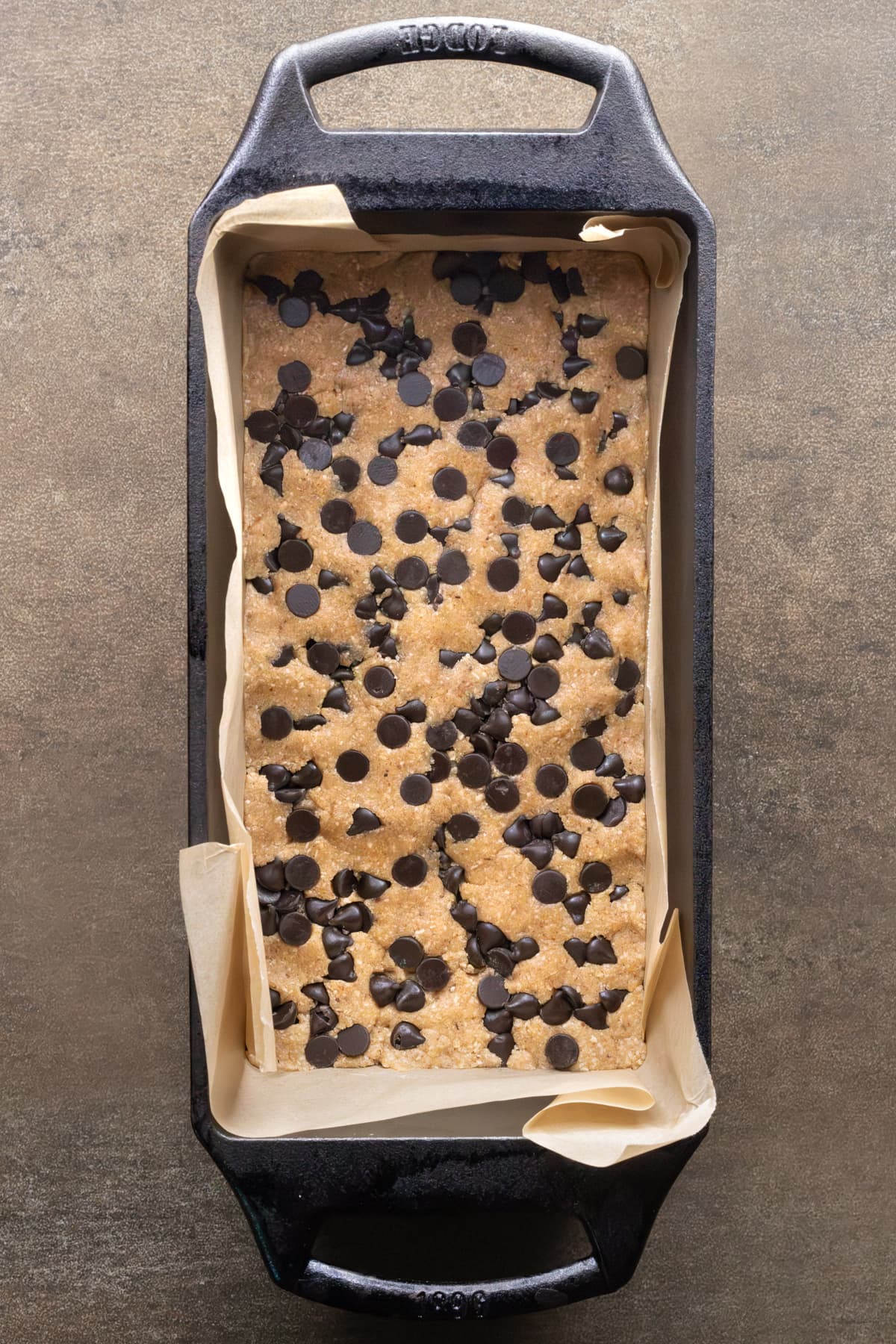 Chewy chocolate chip oat protein bar mixture pressed into a loaf pan, topped with chocolate chips, ready to chill