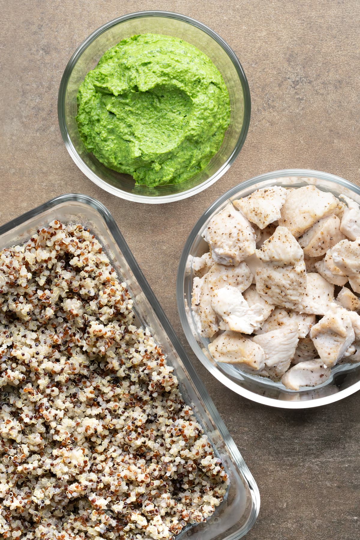 Glass meal prep containers filled with cooked tri-color quinoa, baked chicken pieces and pesto dressing