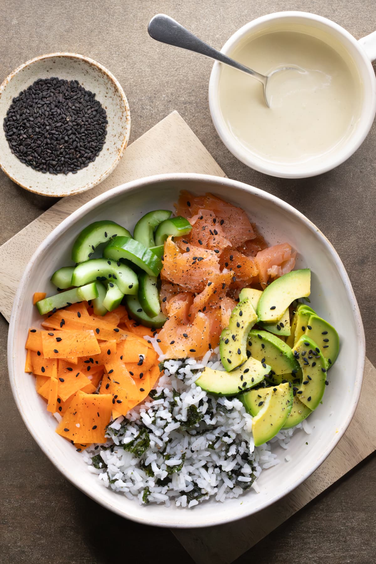 A bowl with smoked salmon, white rice, nori, vegetables, and black sesame seeds, ready for wasabi dressing