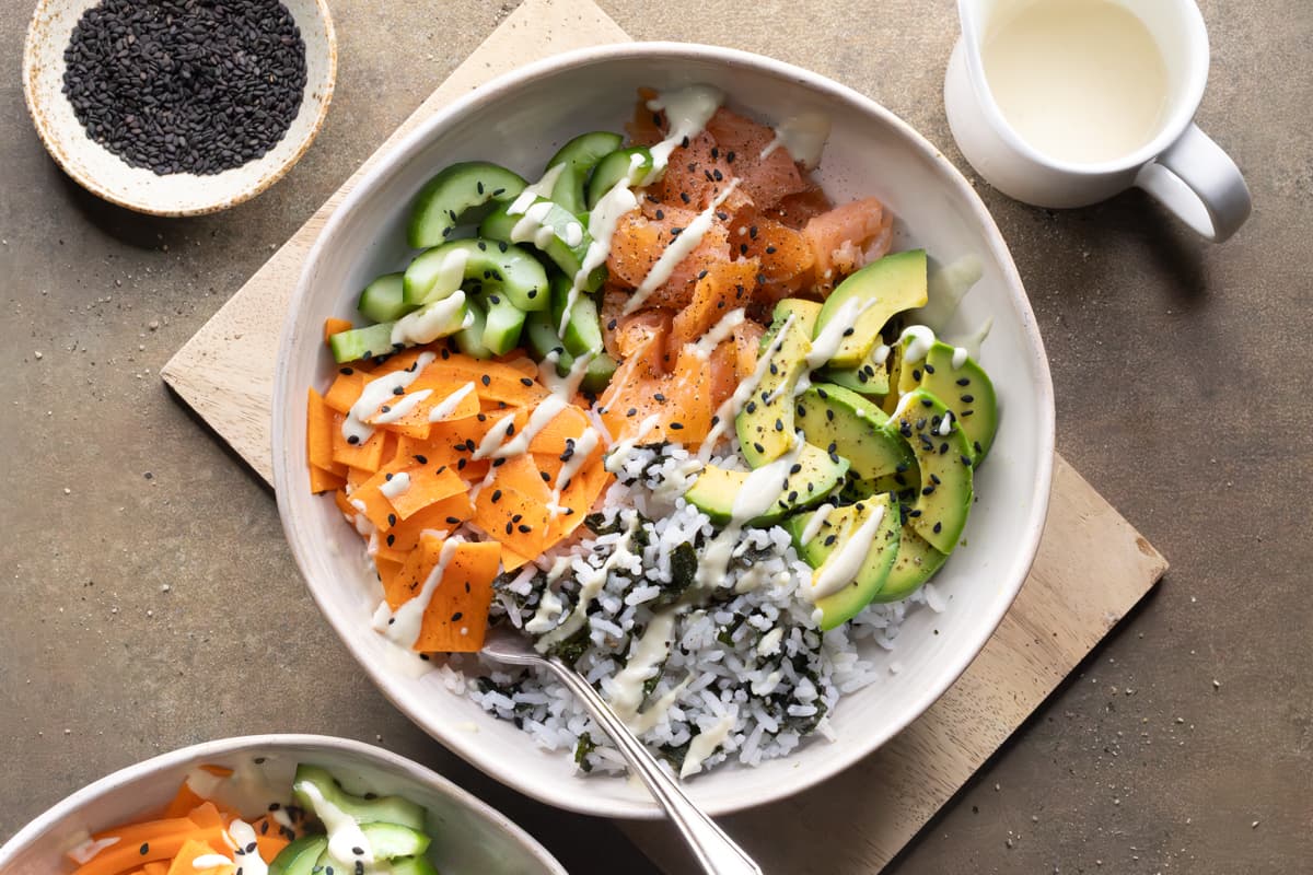 A rice bowl with smoked salmon, vegetables, nori, black sesame seeds, and a drizzle of wasabi dressing