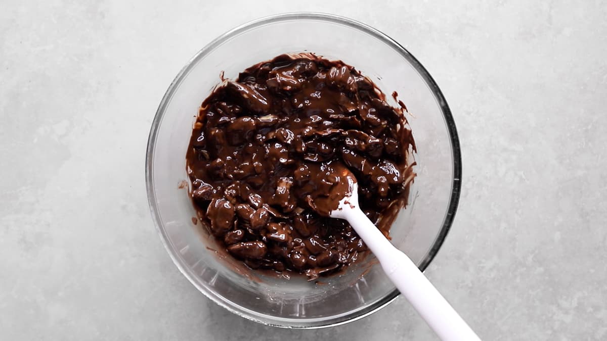 Rocky road mixture of nuts, dried fruit, and coconut folded into melted chocolate in a mixing bowl with a spatula