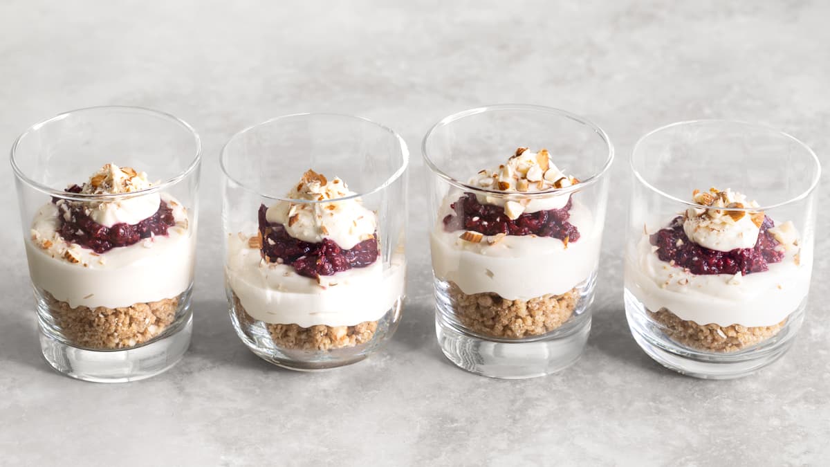 Four no-bake cheesecake cups in glasses topped with chopped almonds and layered with raspberry compote and cheesecake filling