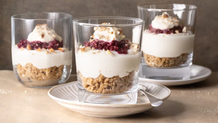Three no-bake cheesecake cups with layers of cheesecake filling, raspberry compote, and oat and almond crumble served in glasses