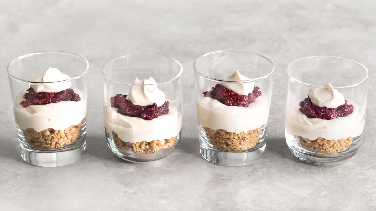 Four assembled no-bake cheesecake cups in glasses with layers of cheesecake filling, raspberry compote, and oat almond crumble