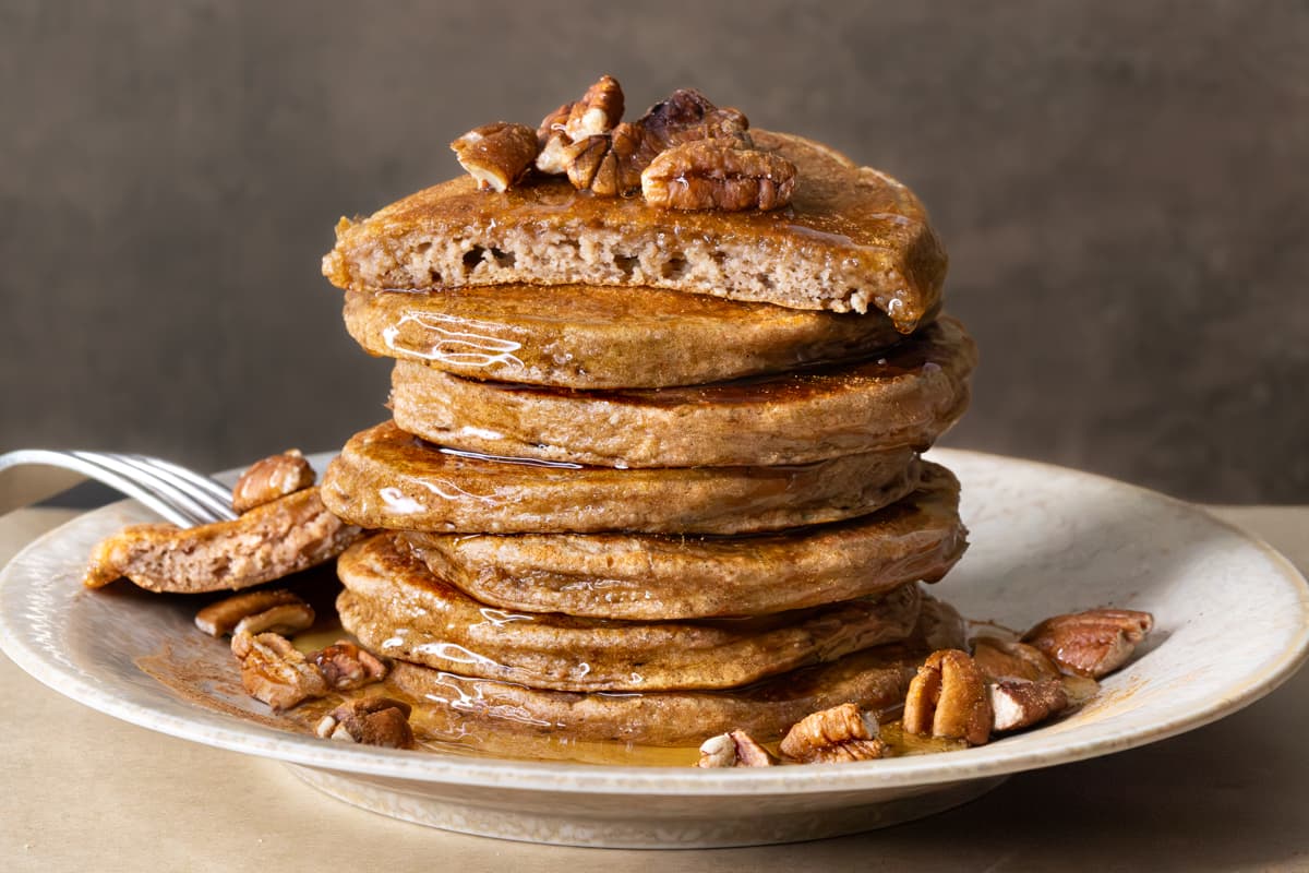 A stack of pancakes with the top pancake cut in half, topped with chopped pecans and brown rice syrup